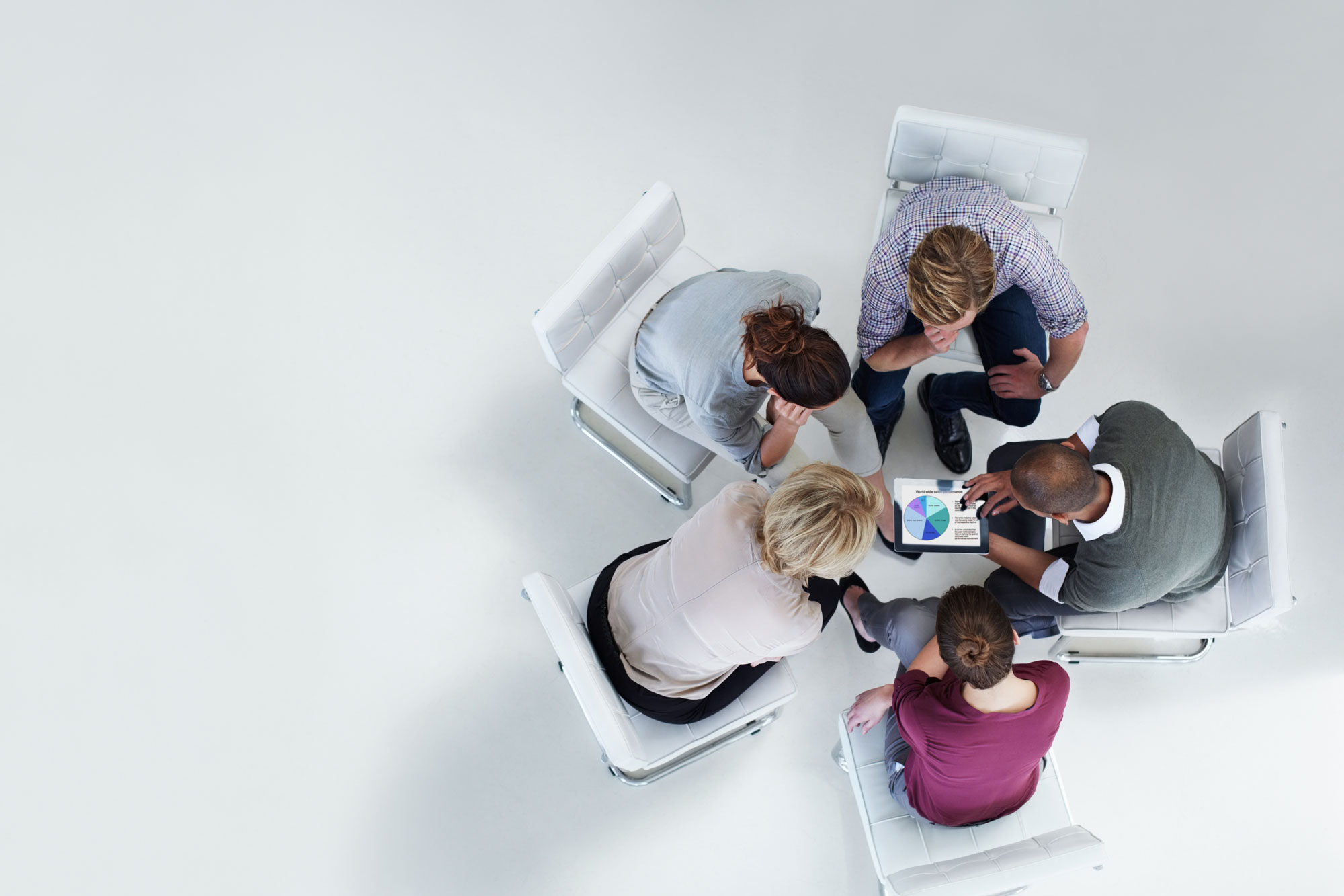 A stock photo of an aerial view of five businesspeople sitting in white chairs and leaning forward to look at a tablet with a pie chart