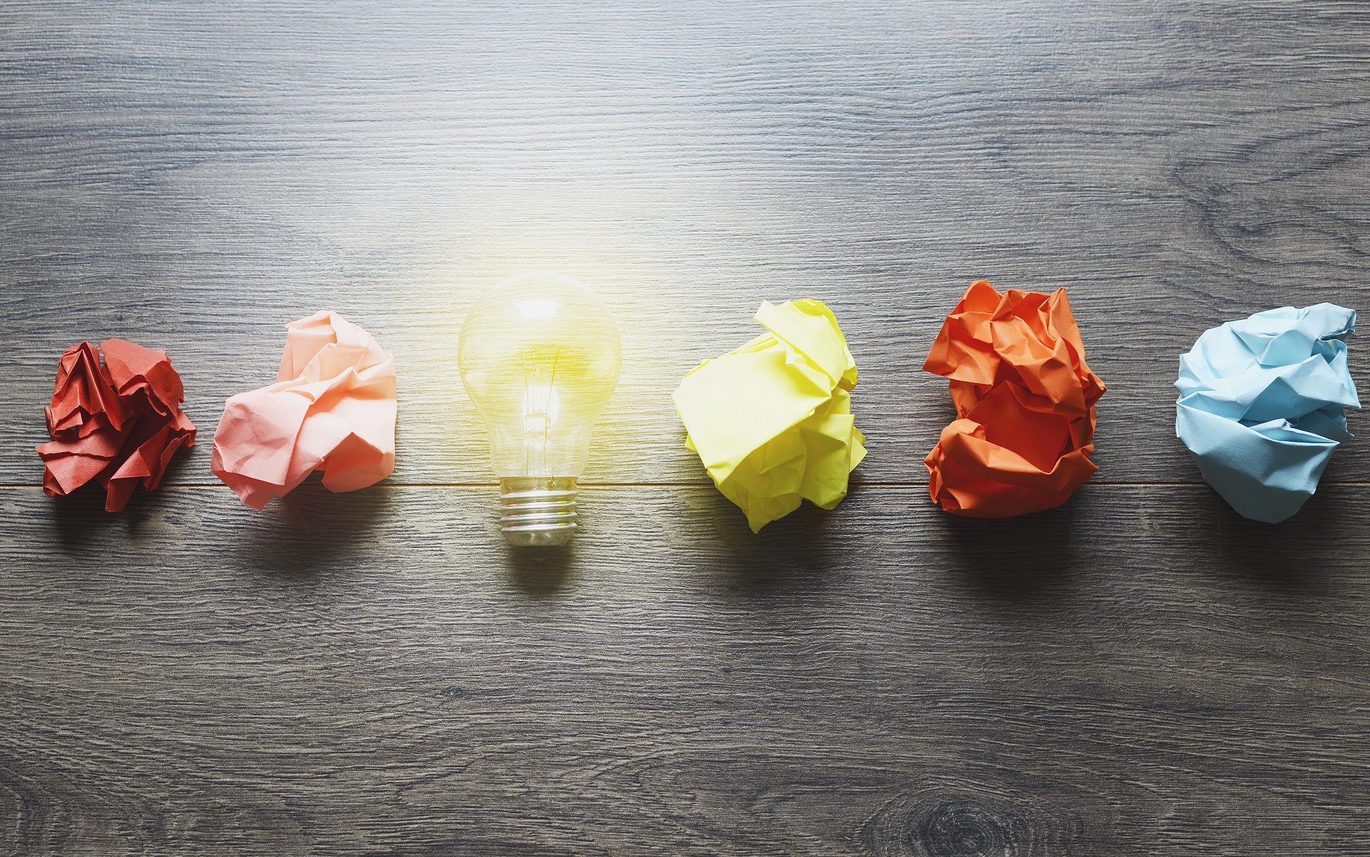 A stock image of crumpled up pieces of multicolored paper in a horizontal line with a lit up lightbulb between two of the pieces of paper